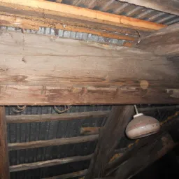 Drewniana konstrukcja stropu poddasza z widocznymi uszkodzeniami, fragmentami blachy falistej i wiszącą starą lampą.