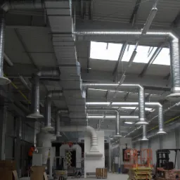 System wentylacji przemysłowej z rurami ze stali nierdzewnej w hali produkcyjnej, widoczne wózki widłowe i elementy maszyn.