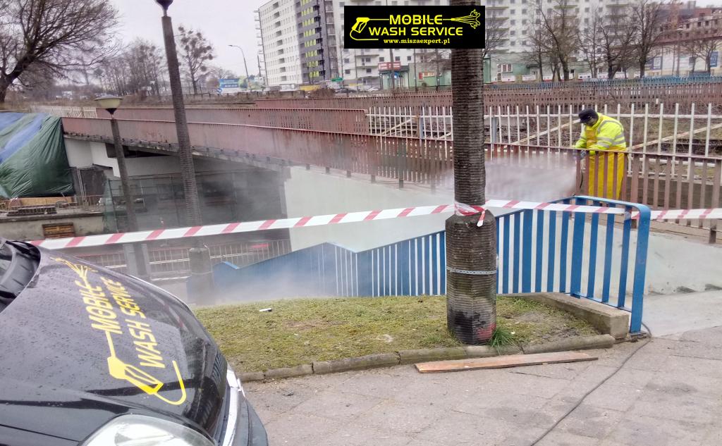 Pracownik w żółtym kombinezonie czyści pod ciśnieniem metalowe elementy wiaduktu nad przejściem podziemnym, widoczny fragment samochodu z logo firmy świadczącej usługi mycia mobilnego.