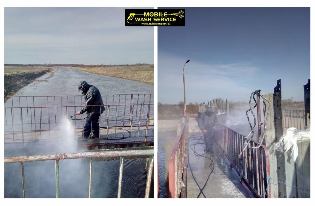 Czyszczenie mostu nad kanałem wodnym za pomocą myjki ciśnieniowej przez pracownika w kombinezonie ochronnym, widoczne strumienie wody pod ciśnieniem i logo firmy na górze kadru.