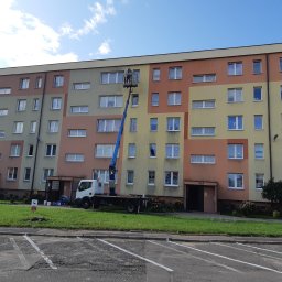 P.U. PRZEMYSŁAW MURAWSKI - Pracownik na podnośniku koszowym czyści elewację bloku. Widoczny fragment budynku z oknami, trawnik i zaparkowany samochód. Słoneczny dzień.