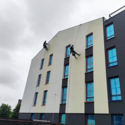 P.U. PRZEMYSŁAW MURAWSKI - Dwóch alpinistów maluje elewację bloku. Okna zabezpieczone folią. Część ściany pomalowana na szaro, część na biało. Pochmurne niebo w tle.
