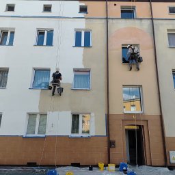 P.U. PRZEMYSŁAW MURAWSKI - Dwóch alpinistów maluje elewację bloku. Częściowo pomalowana ściana, widoczne zabezpieczenia okien folią i pojemniki z farbą na dole. Prace na wysokościach.