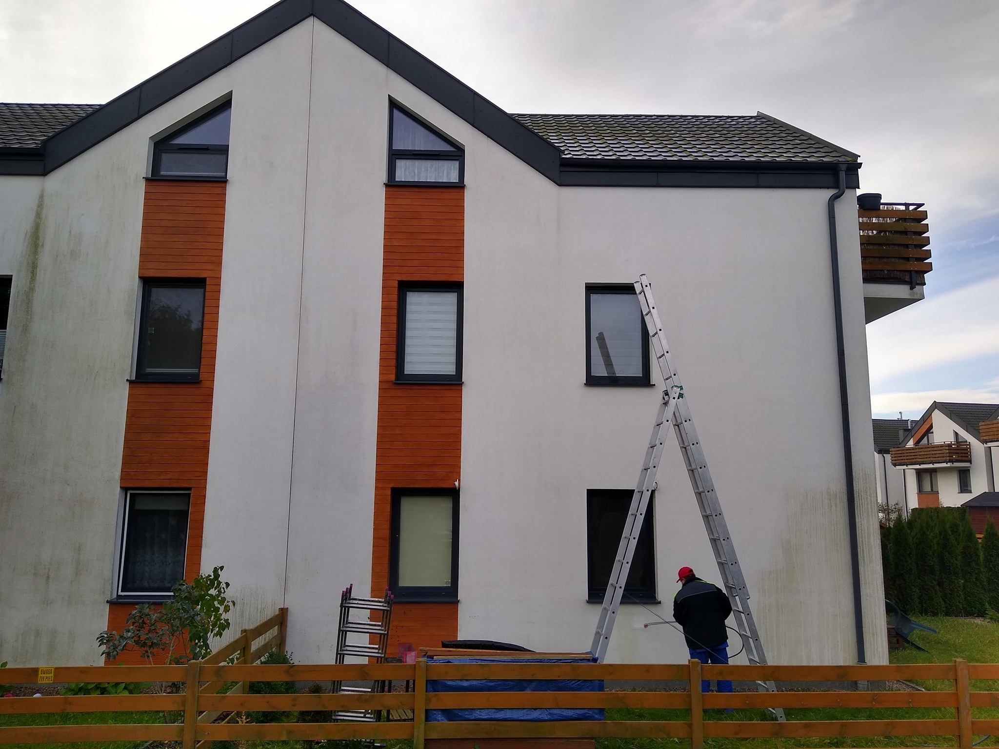 Mycie elewacji domu z widocznymi zabrudzeniami. Pracownik na drabinie używa myjki ciśnieniowej. Widoczne efekty czyszczenia.