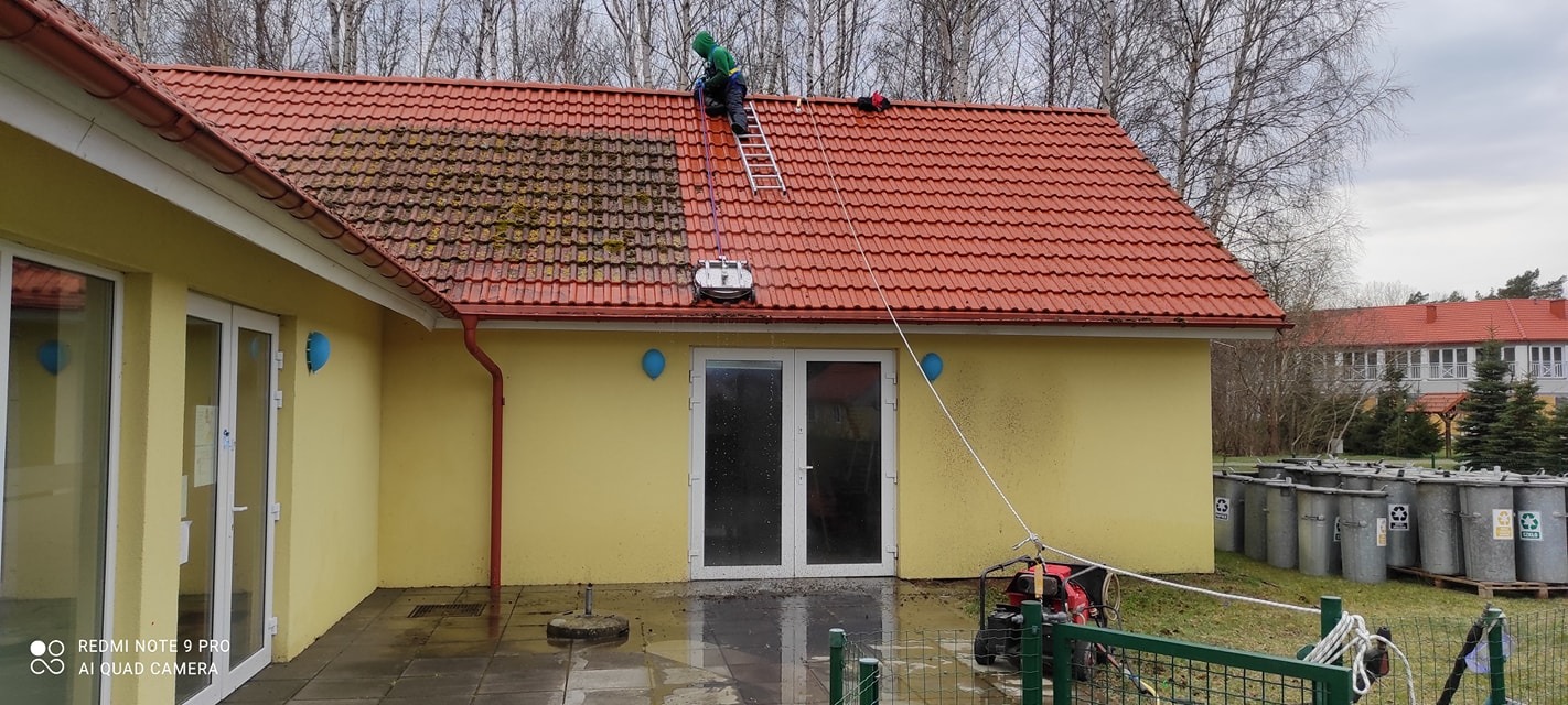 Czyszczenie dachu z czerwonej dachówki myjką ciśnieniową. Widoczna różnica między umytą a zabrudzoną częścią dachu. Pracownik na drabinie, żółty budynek w tle.