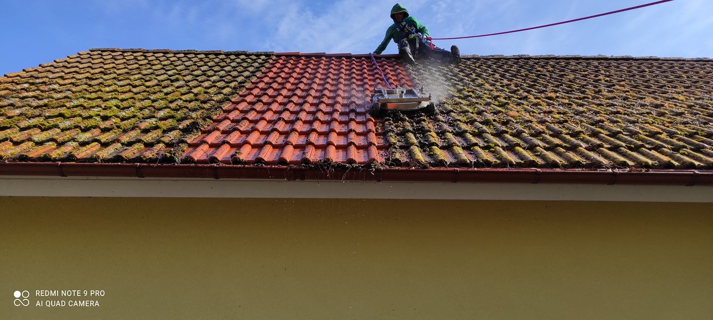 Czyszczenie dachu z mchu i zabrudzeń za pomocą myjki ciśnieniowej. Widoczna różnica między czystą a zabrudzoną częścią dachu. Praca na wysokościach z zabezpieczeniami.