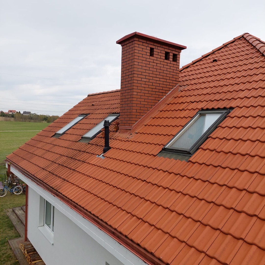 Czerwony dach z dachówką ceramiczną, komin z cegły i okna dachowe. Widok z góry na dom z ogrodem w tle. Charakterystyczny kształt dachówki.