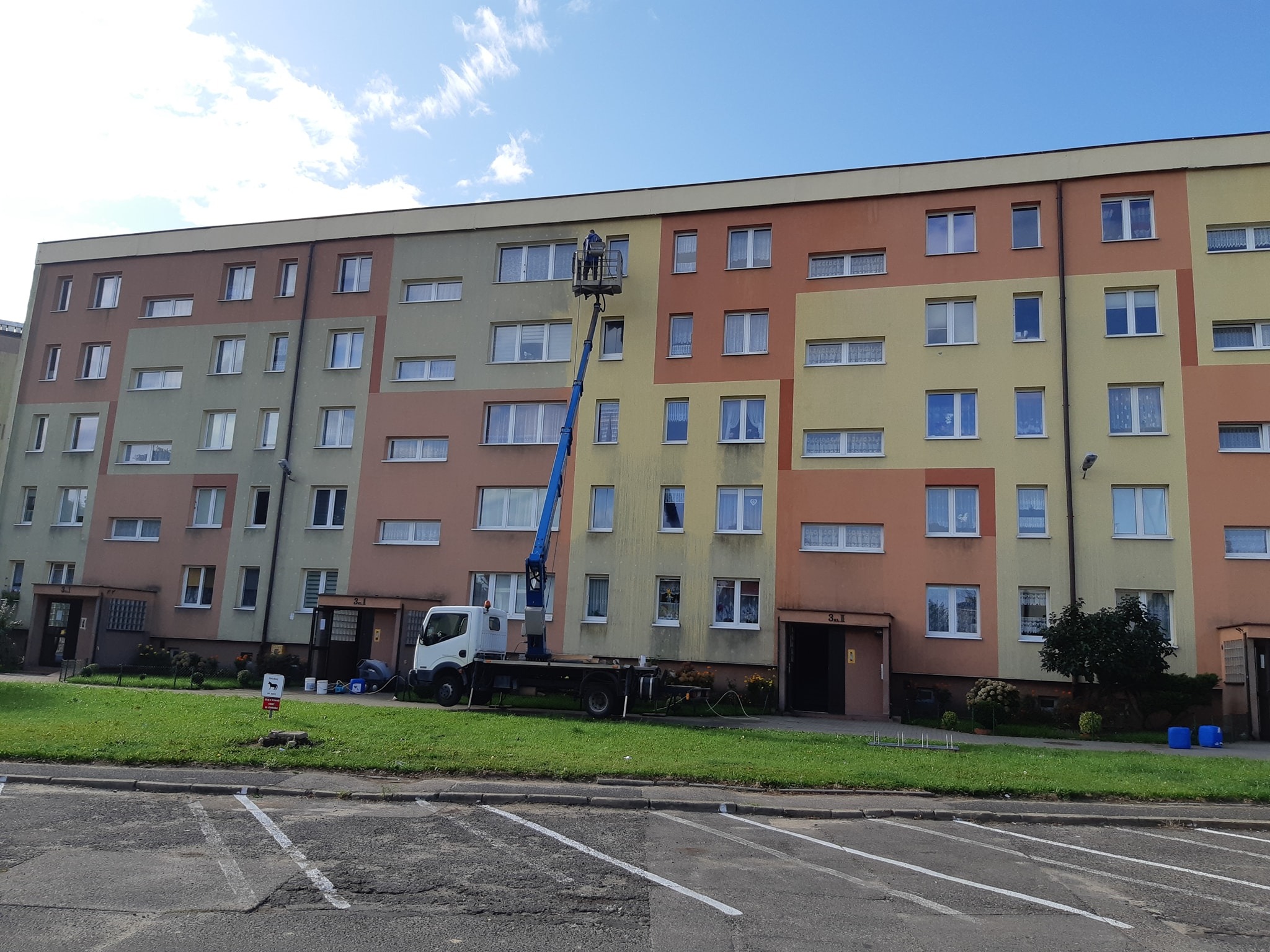 Pracownik na podnośniku koszowym czyści elewację bloku. Widoczny fragment budynku z oknami, trawnik i zaparkowany samochód. Słoneczny dzień.