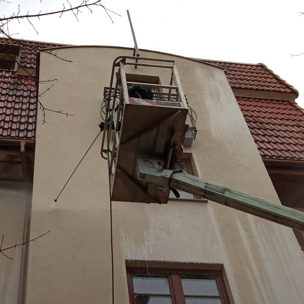 Mycie elewacji budynku z podnośnika koszowego. Strumień wody usuwa zabrudzenia, odsłaniając czystą powierzchnię. Widoczny fragment dachu z czerwoną dachówką.