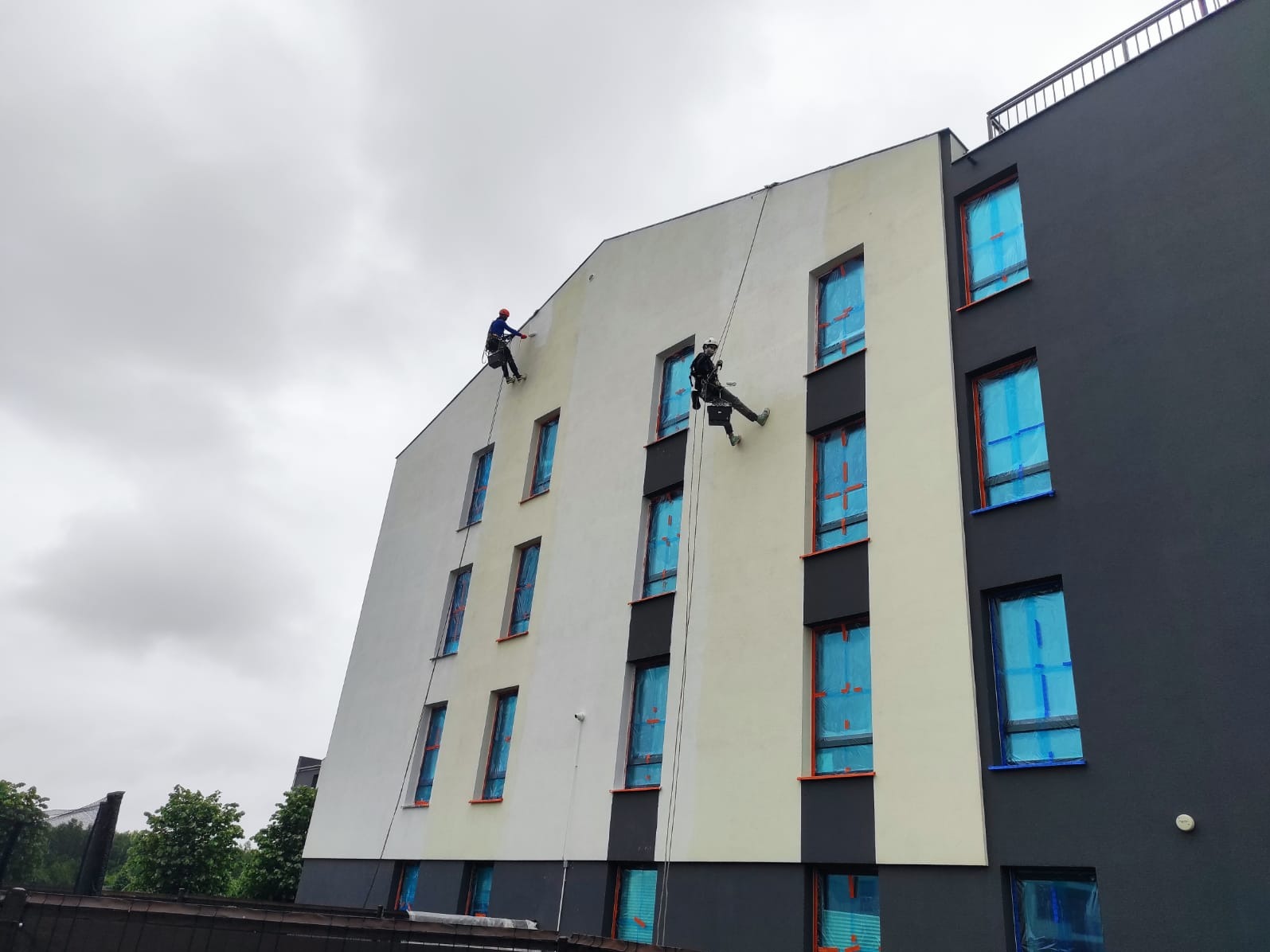 Dwóch alpinistów maluje elewację bloku. Okna zabezpieczone folią. Część ściany pomalowana na szaro, część na biało. Pochmurne niebo w tle.