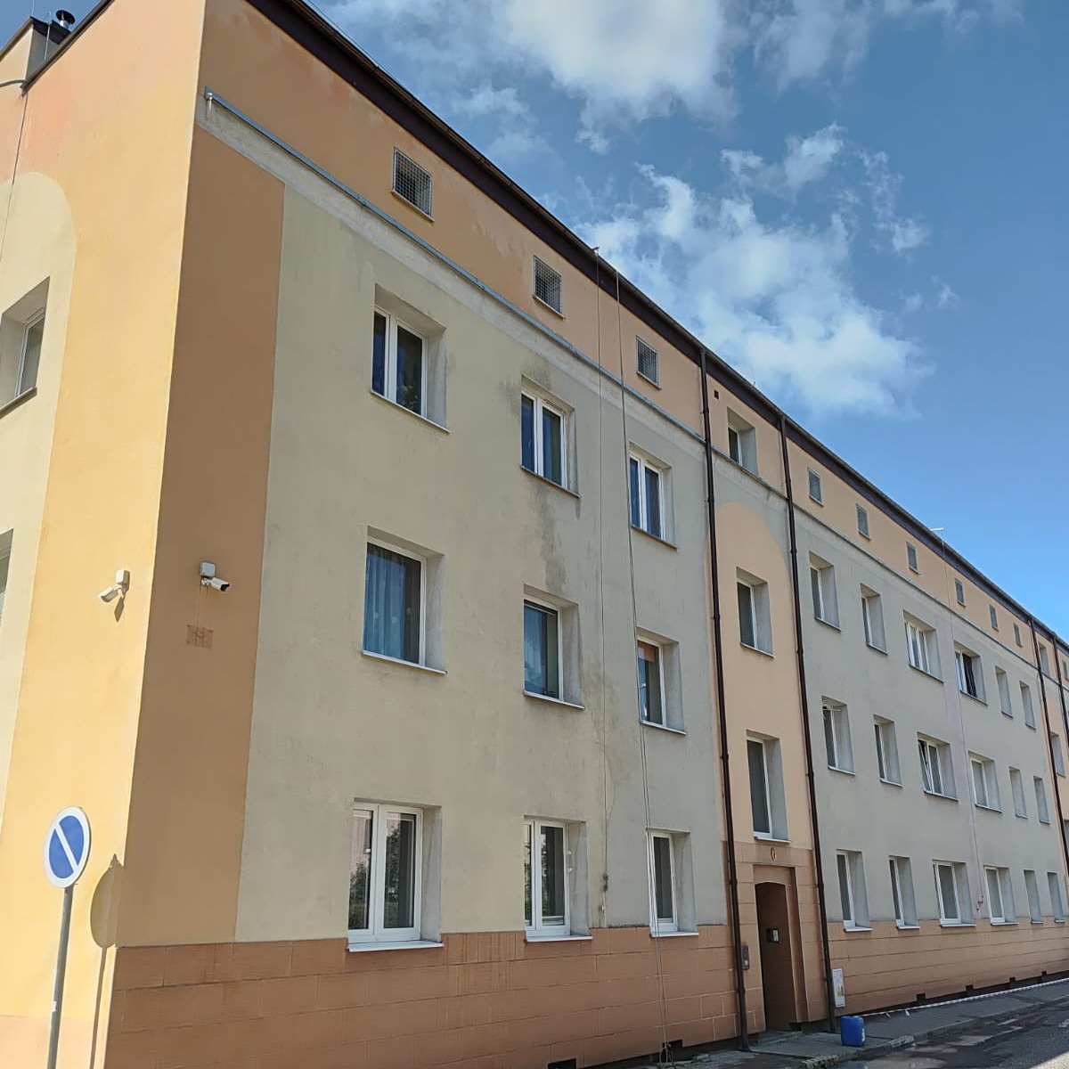 Odnowiona elewacja bloku mieszkalnego w odcieniach beżu i brązu, widok z dołu, błękitne niebo w tle. Widoczne okna z białymi ramami i elementy dekoracyjne.