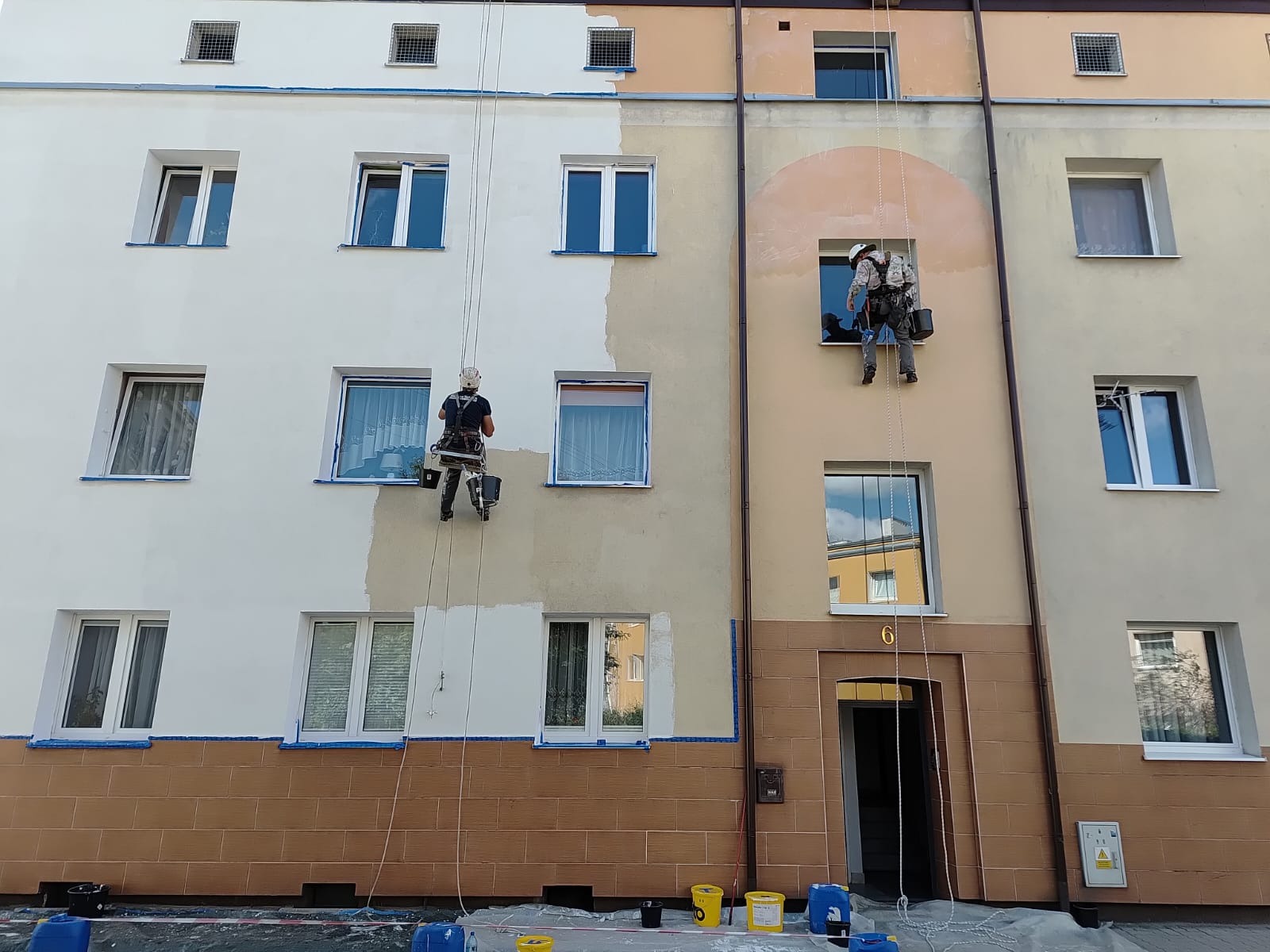 Dwóch alpinistów maluje elewację bloku. Częściowo pomalowana ściana, widoczne zabezpieczenia okien folią i pojemniki z farbą na dole. Prace na wysokościach.