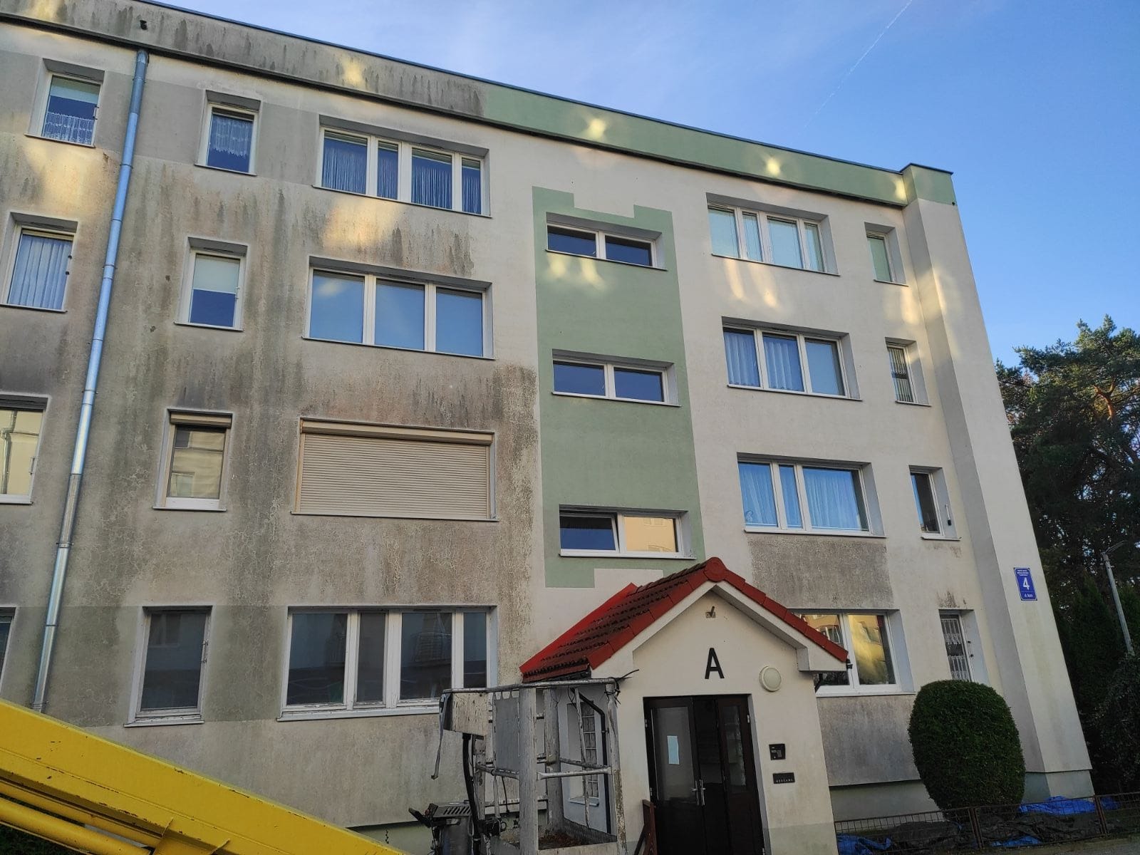 Brudna elewacja bloku przed czyszczeniem, widoczny podnośnik koszowy. Część fasady z wyraźnymi zabrudzeniami, okna z roletami. Wejście do budynku z daszkiem.