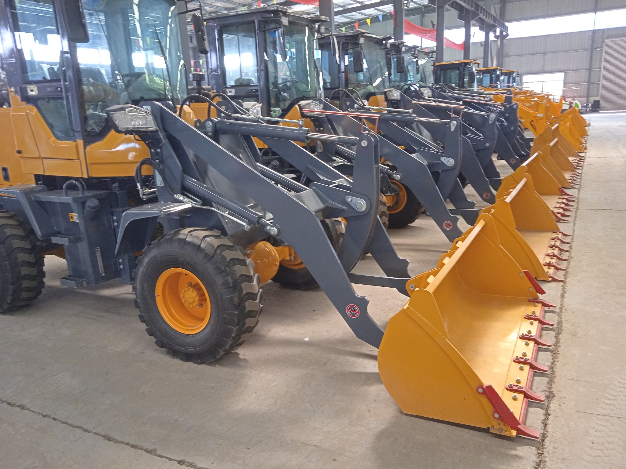 Rząd nowych, żółto-szarych ładowarek kołowych z zębami na łyżkach, ustawionych w hali produkcyjnej.