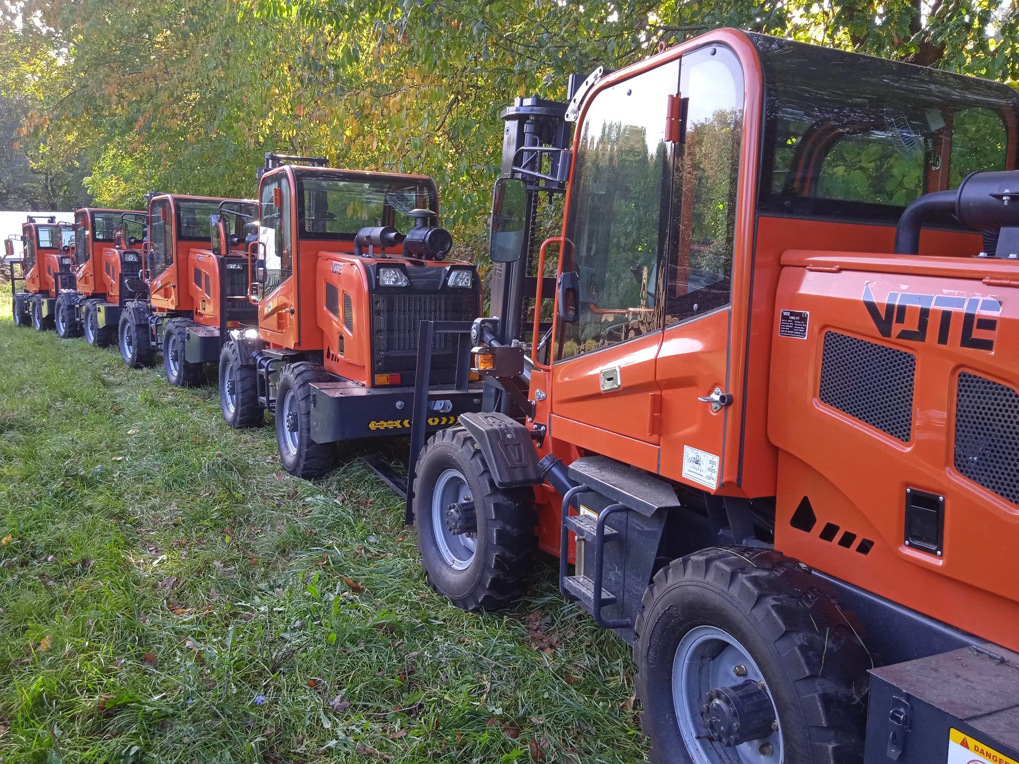 Szereg pomarańczowych, małych ciągników leśnych stoi w Żarnowie na trawiastym terenie pod drzewami, czekając na transport lub użycie.