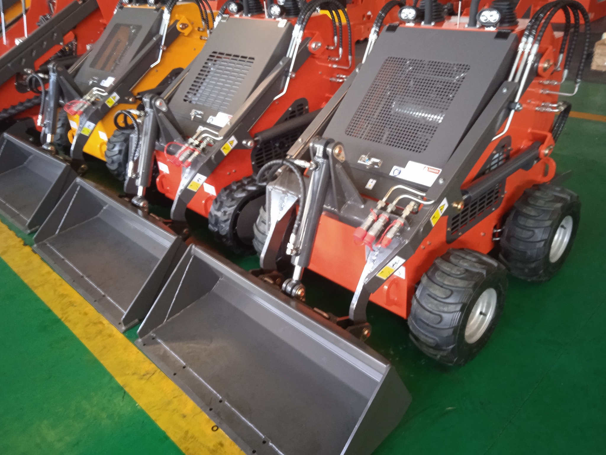 Rząd nowych, pomarańczowych miniładowarek z szarymi osłonami i metalowymi łyżkami na zielono-żółtej podłodze w hali produkcyjnej.