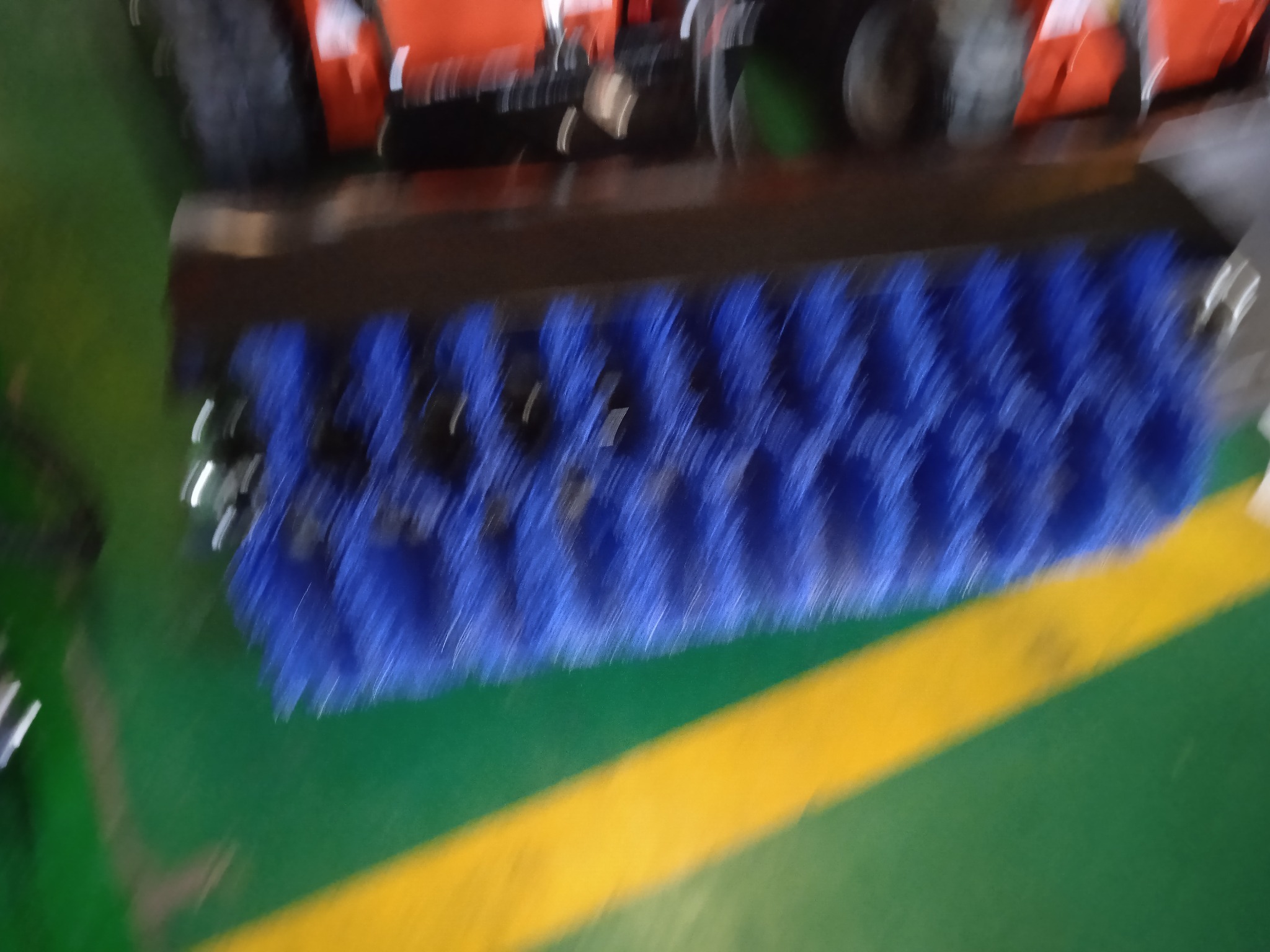 Szczotka zamiatająca z niebieskim włosiem zamontowana na pomarańczowym pojeździe czyszczącym, widoczna na zielonej powierzchni z żółtym pasem.