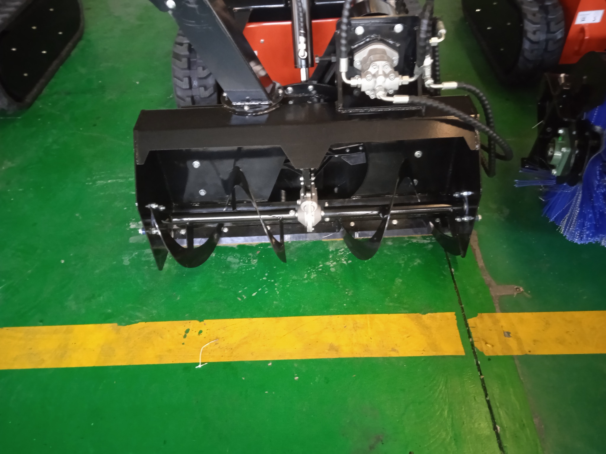 Czarny frez do odśnieżania zamontowany na małej ładowarce gąsienicowej, widok z przodu na zielonej posadzce z żółtą linią.