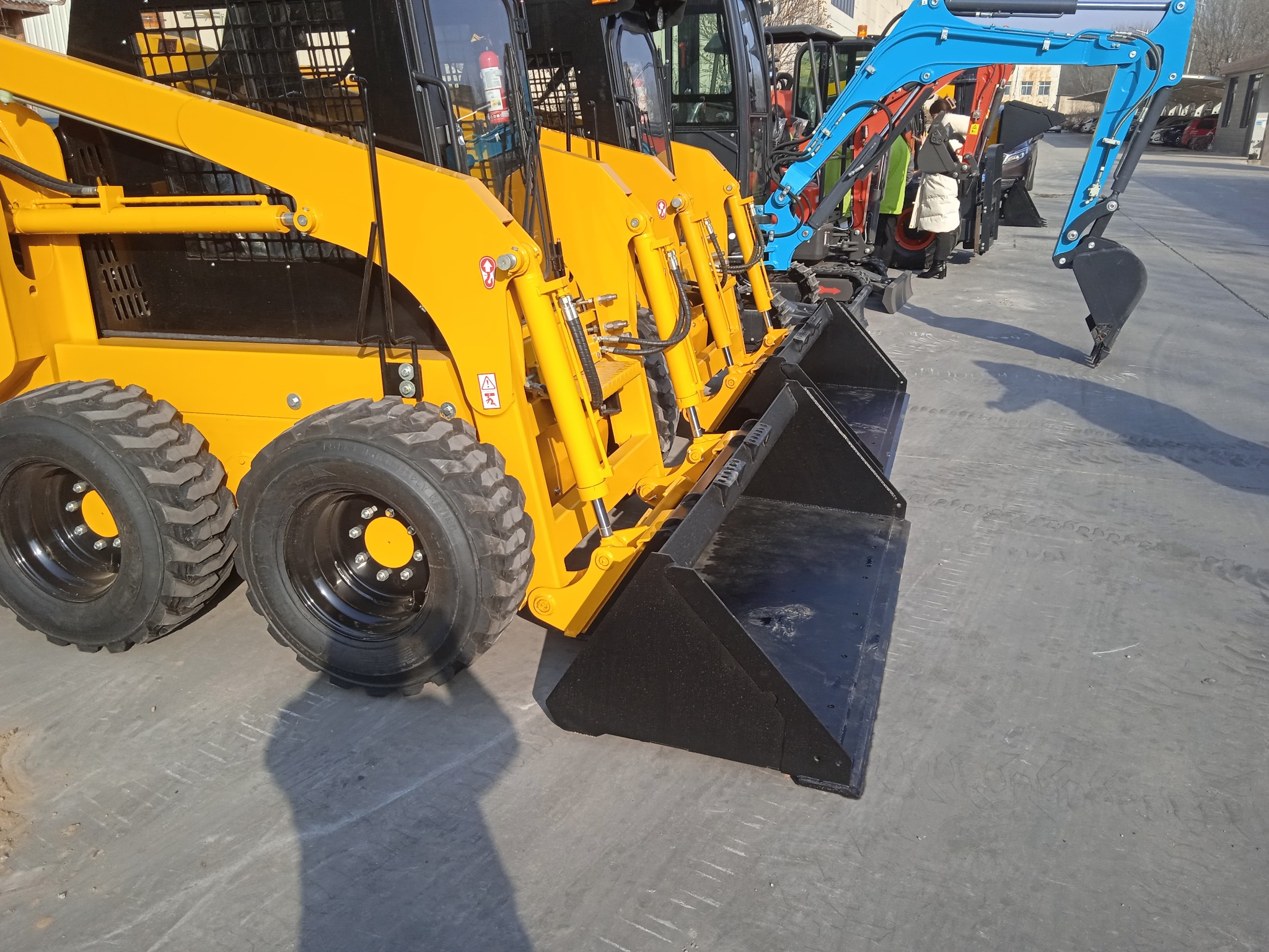 Rząd żółtych miniładowarek ze standardowymi łyżkami, z niebieską minikoparką w tle, prezentowany na placu składowym z betonową nawierzchnią.