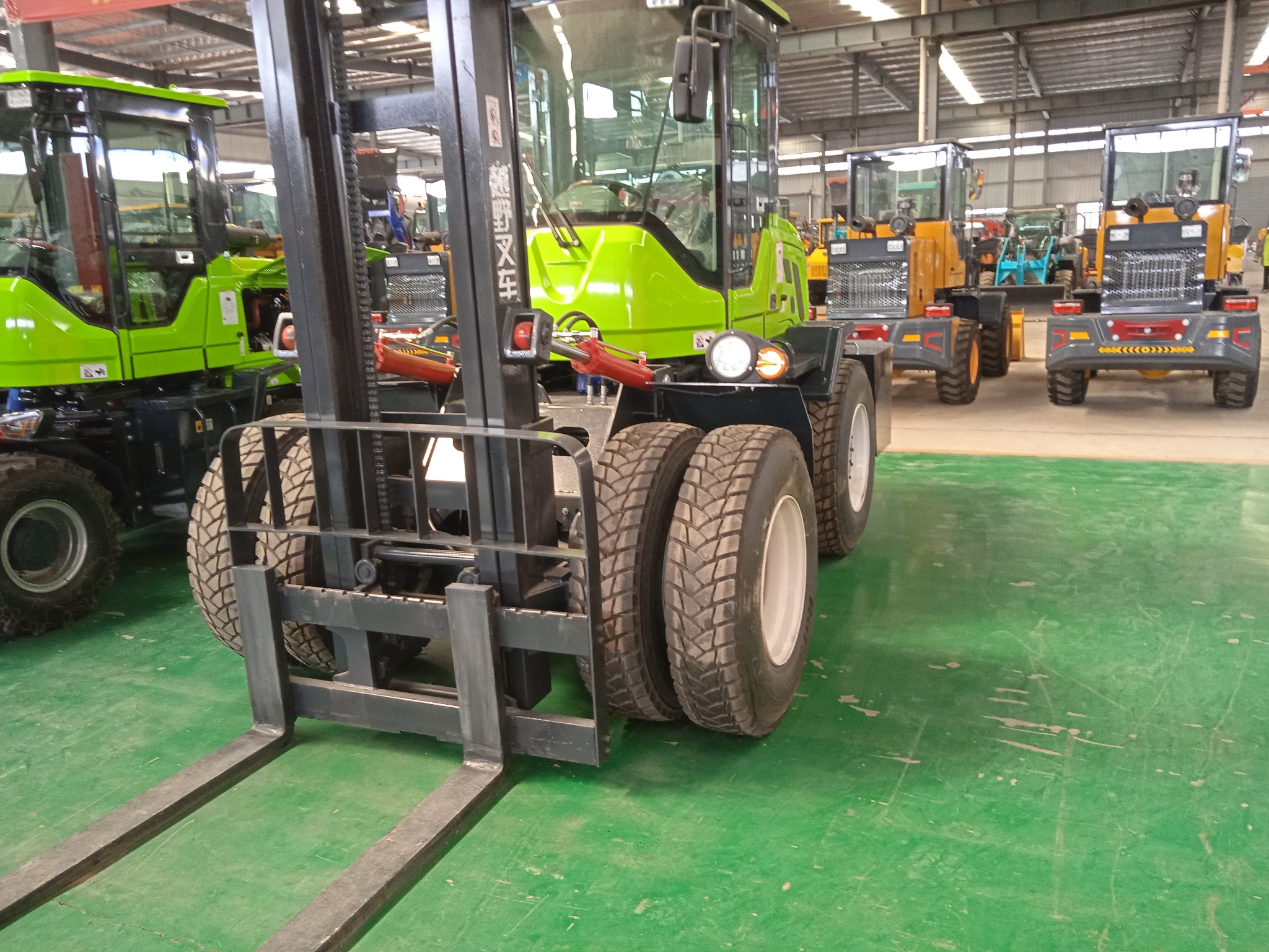 Zielony wózek widłowy z podwójnymi kołami z przodu, widok z bliska na widły i opony, w tle inne maszyny w hali.