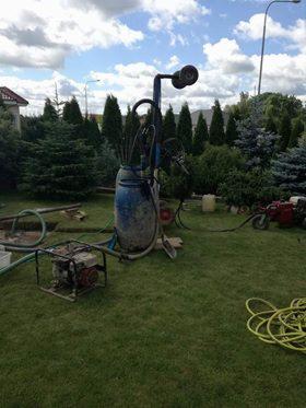 Sprzęt do wiercenia studni głębinowej: niebieska beczka, pompa, węże i generator na trawniku przed domem z widocznymi drzewami iglastymi w tle.