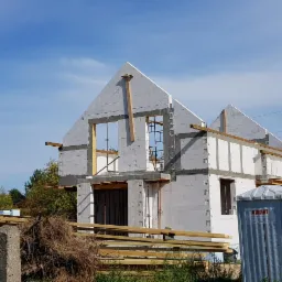 Wznoszenie ścian z białych bloczków na placu budowy, widoczne drewniane elementy konstrukcyjne i przenośna toaleta.