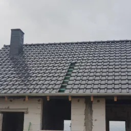 Niedokończony dom z czarnym dachem z dachówki ceramicznej i kominem z tego samego materiału, widoczne fragmenty konstrukcji dachu z zieloną membraną, budynek w stanie surowym otwartym z białego...