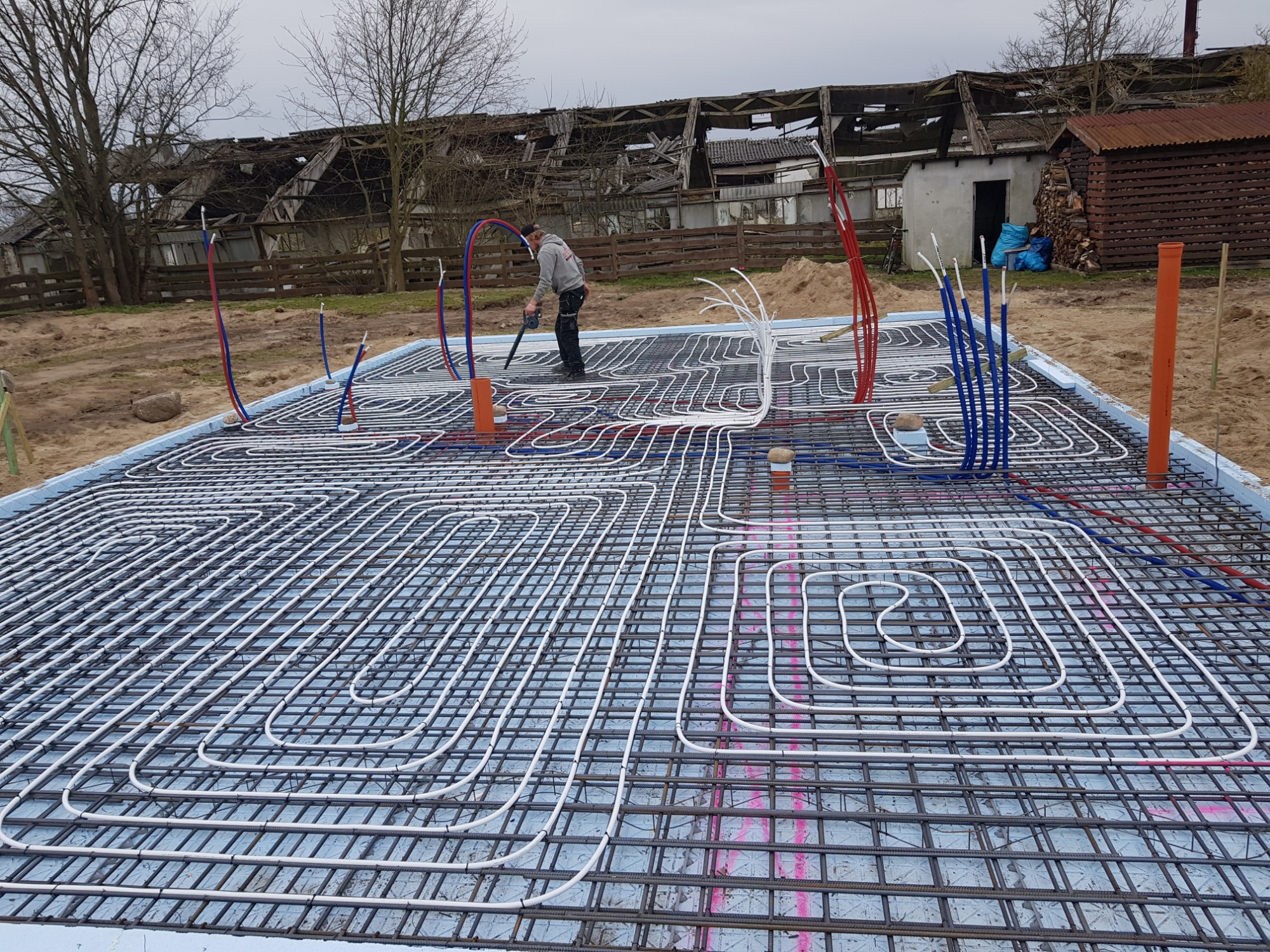 Instalacja ogrzewania podłogowego w fundamencie, widoczne białe rury w siatce zbrojeniowej, z pracownikiem używającym dmuchawy do czyszczenia, w tle zrujnowany budynek gospodarczy.