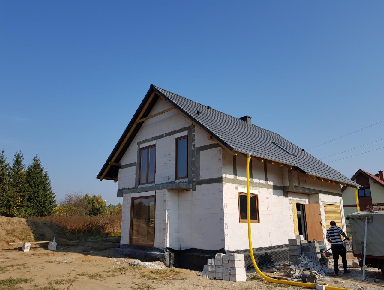 Wznoszony dom murowany z białych bloczków, z zamontowanymi oknami z brązowymi ramami, pokryty szarą dachówką, widoczny pracownik i żółty wąż doprowadzający wodę.