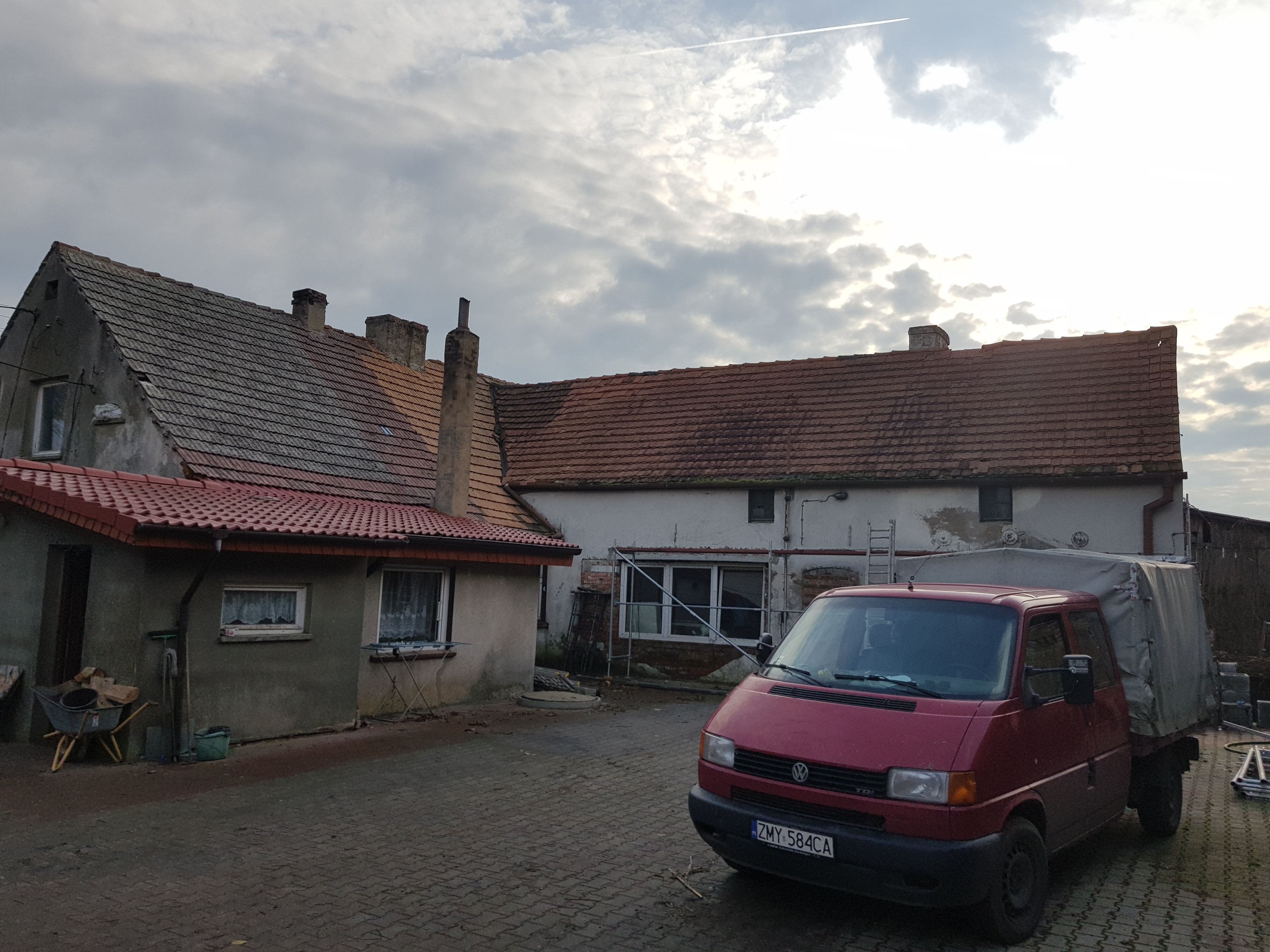 Starszy dom murowany z częściowo odnowionym dachem, widoczny czerwony bus transportowy i rozstawiona drabina sugerują prace remontowe.