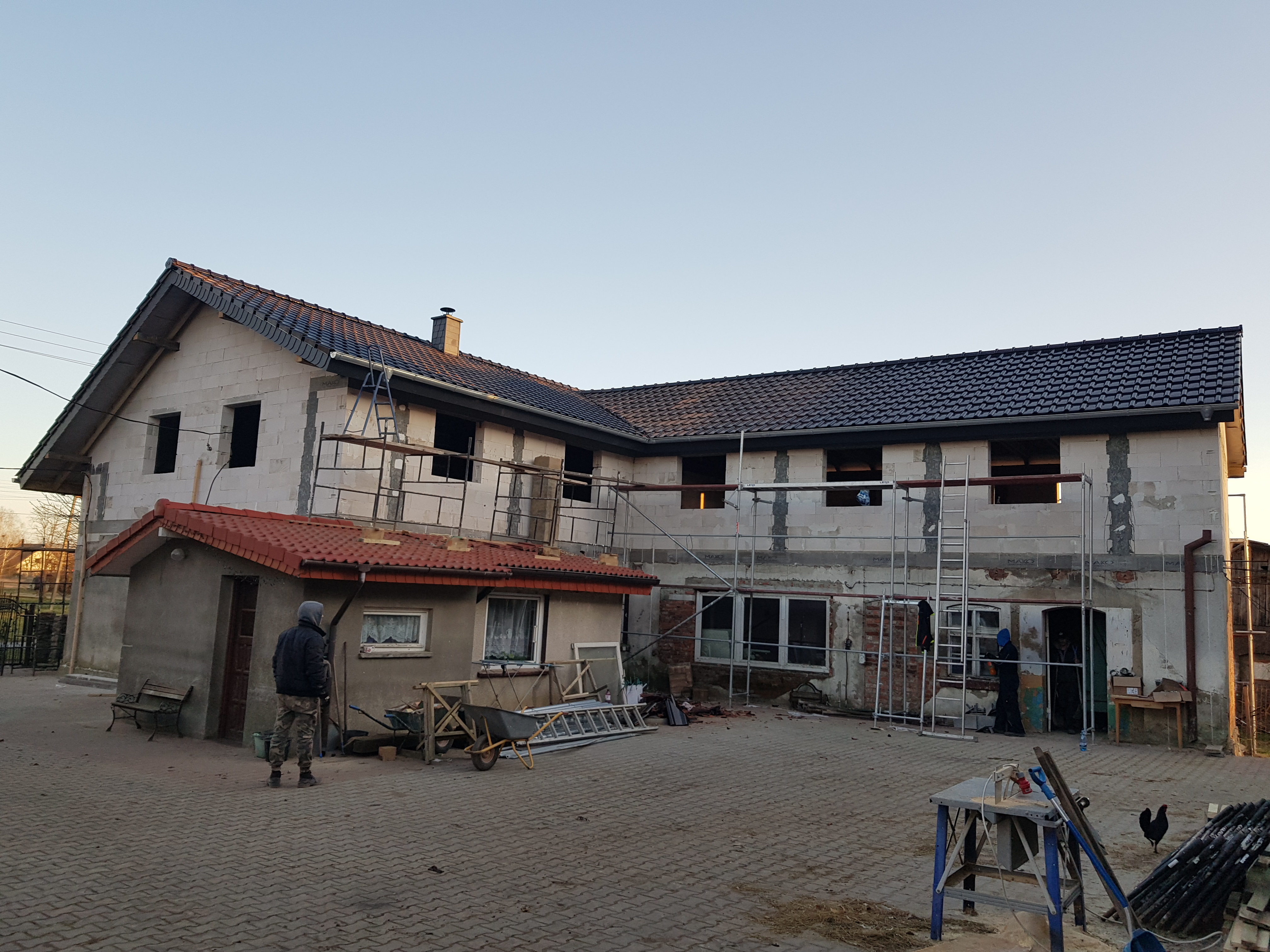 Budowa domu z białego pustaka w Wierzbówku, widoczne rusztowania, nowy dach z czarnej dachówki oraz stary budynek z czerwoną dachówką, na podwórku pracownicy i narzędzia.