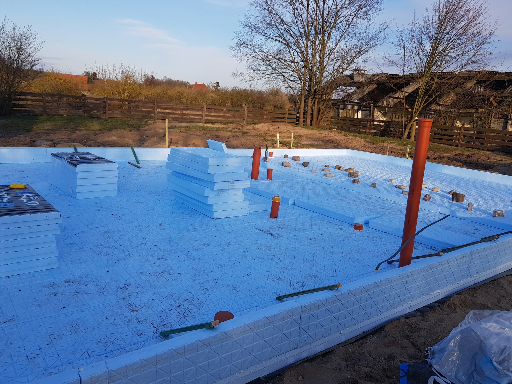 Przygotowanie fundamentów pod dom: płyta fundamentowa z izolacją termiczną z polistyrenu ekstrudowanego, widoczne stosy płyt izolacyjnych i pionowe rury instalacyjne w wykopie, w tle drewniany płot...