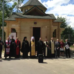 Grupa osób, w tym duchowni w strojach liturgicznych, przed nowo wybudowaną, małą drewnianą kaplicą z zielonym dachem w słoneczny dzień.