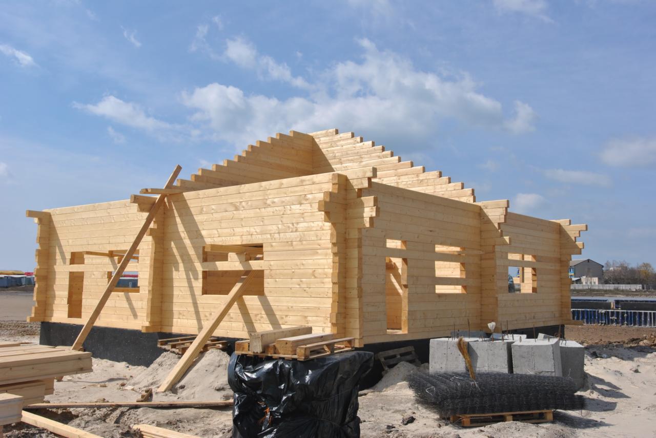 Surowy drewniany szkielet domu w trakcie budowy, z widocznymi belkami stropowymi i otworami okiennymi, na tle błękitnego nieba z chmurami, z paletami i materiałami budowlanymi na pierwszym planie.