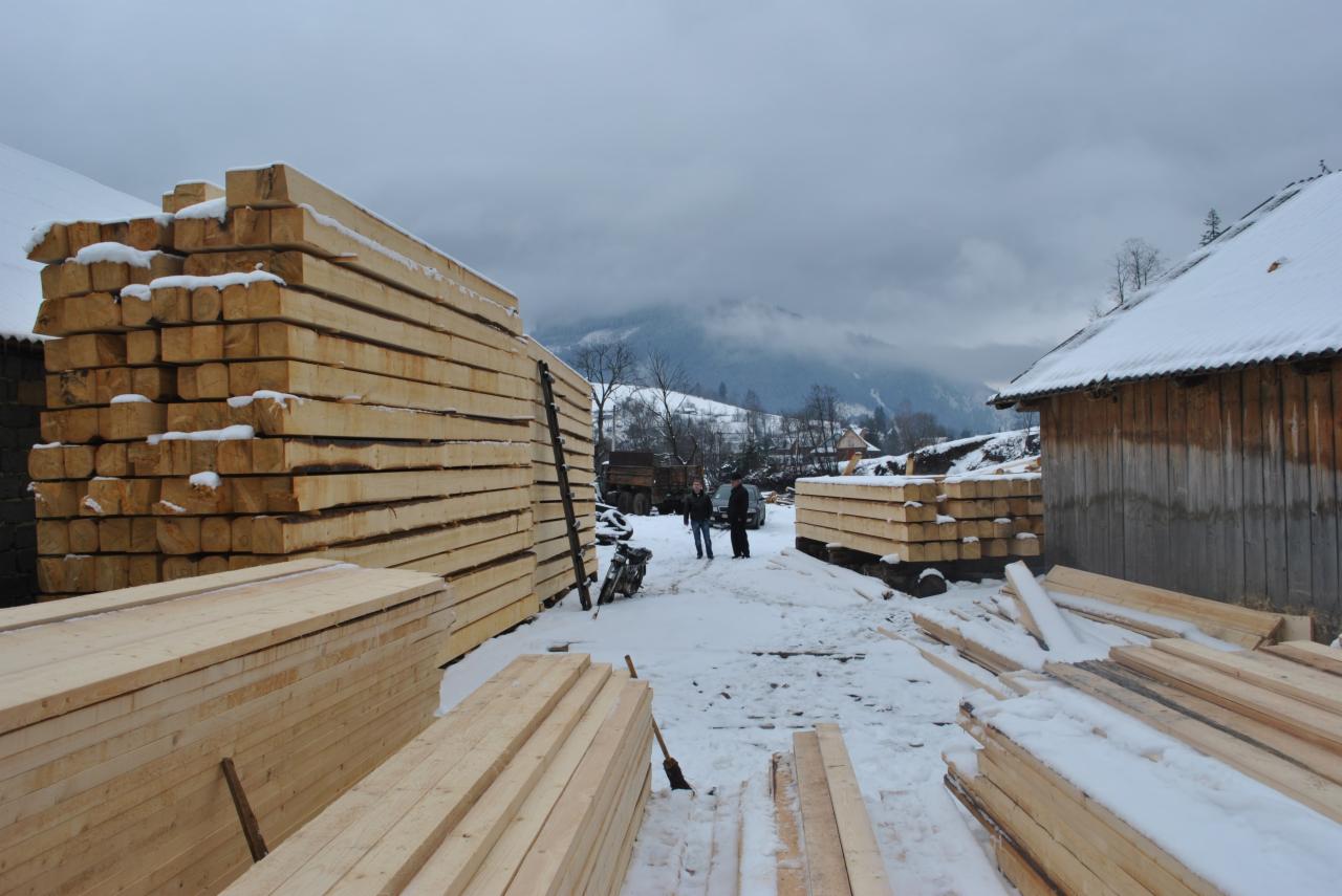 Stosy świeżo obrobionego drewna konstrukcyjnego, pokryte śniegiem, ułożone na placu budowy z widokiem na zalesione wzgórza w tle.