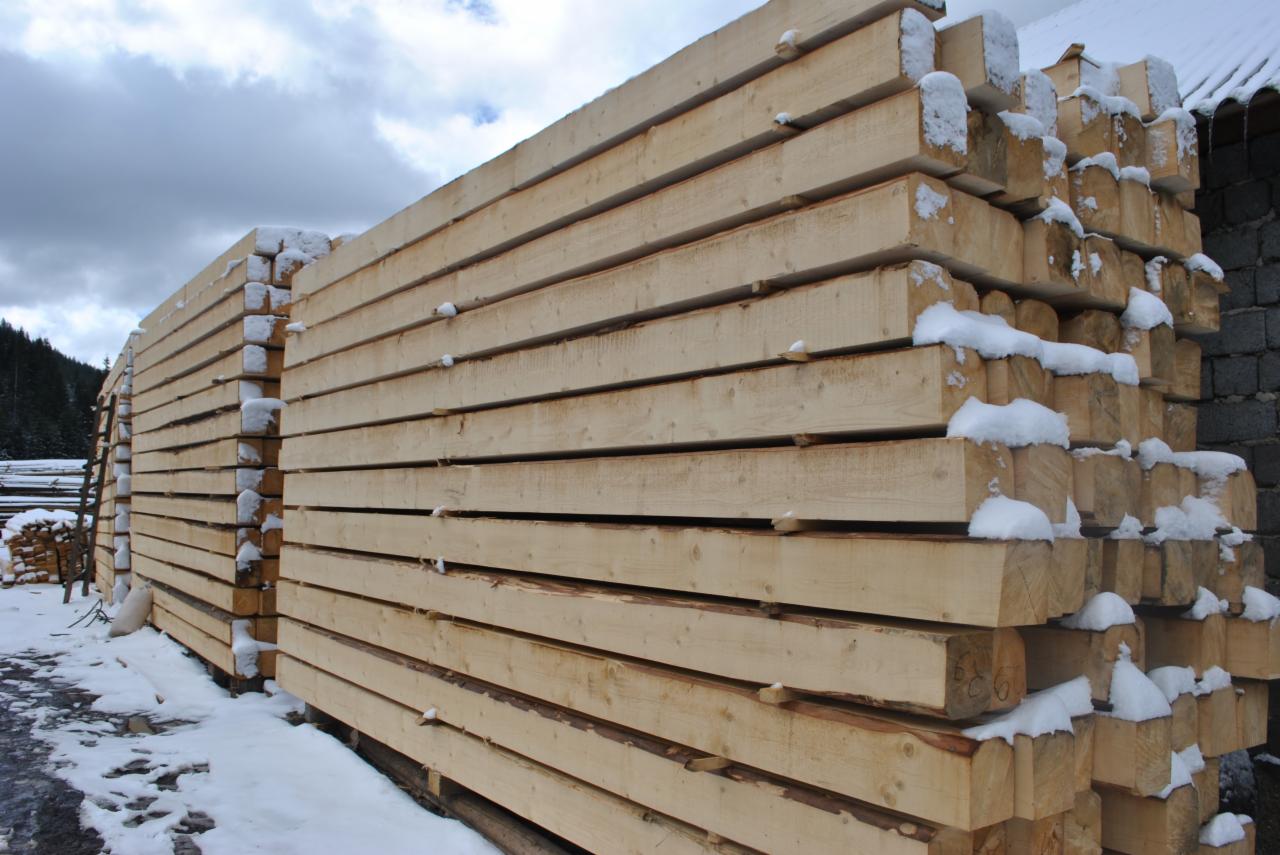 Stos belek z jasnego drewna, pokrytych śniegiem, ułożonych w rzędy, z widocznym fragmentem ściany budynku w tle.
