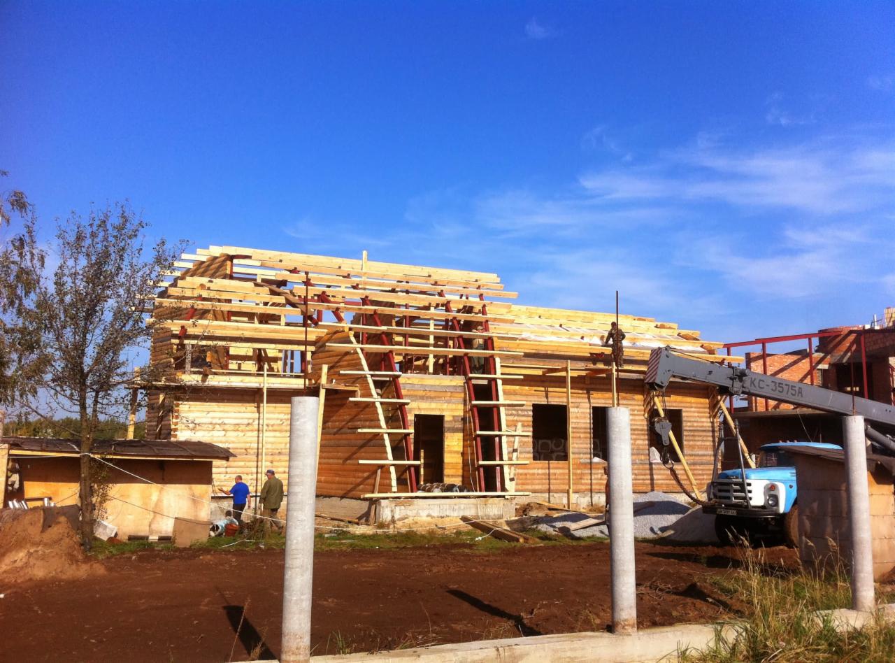 Budowa domu z bali w trakcie realizacji, widoczny dźwig budowlany, drewniana konstrukcja dachu i ściany z bali, dwóch pracowników w oddali, błękitne niebo.