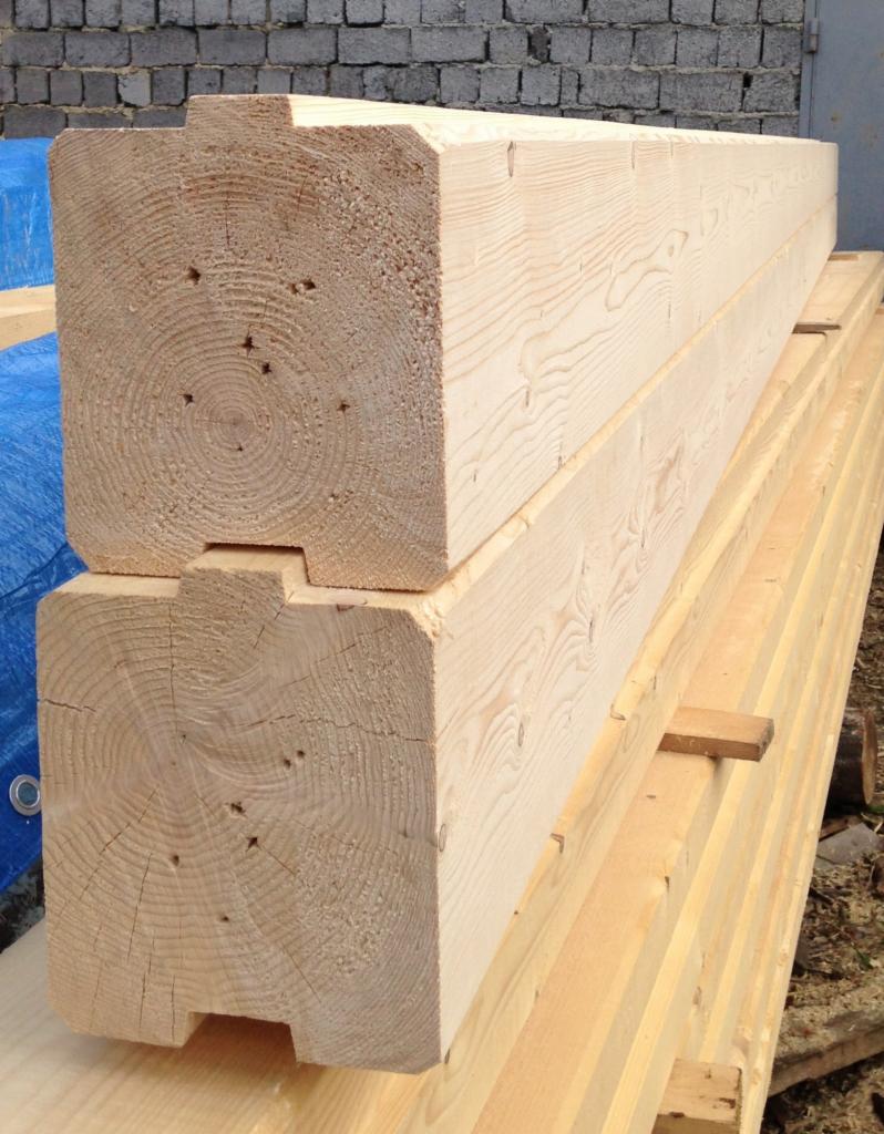 Dwa bale z jasnego drewna, z wyfrezowanymi rowkami, ułożone jeden na drugim na stosie desek, z widocznymi słojami i małymi otworami, w tle mur z szarych cegieł i fragment niebieskiej plandeki.