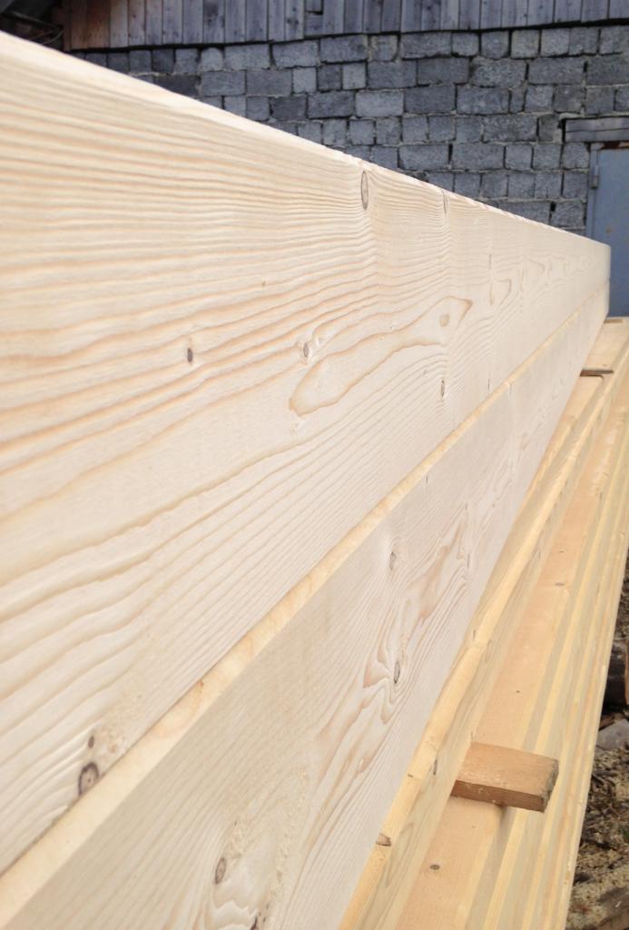 Stos świeżo obrobionych, jasnych belek drewnianych z wyraźnym rysunkiem słojów, przygotowanych do budowy domu z bali, na tle szarego muru z cegieł.