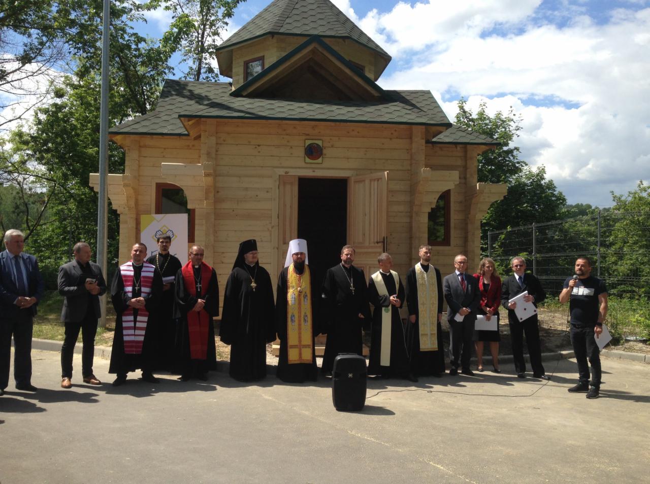 Grupa osób, w tym duchowni w strojach liturgicznych, przed nowo wybudowaną, małą drewnianą kaplicą z zielonym dachem w słoneczny dzień.