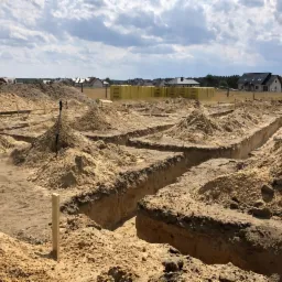 Przygotowanie terenu pod budowę domu: wykopane rowy fundamentowe w piaszczystej glebie, widoczne żółte bloczki budowlane w tle, częściowo zachmurzone niebo.