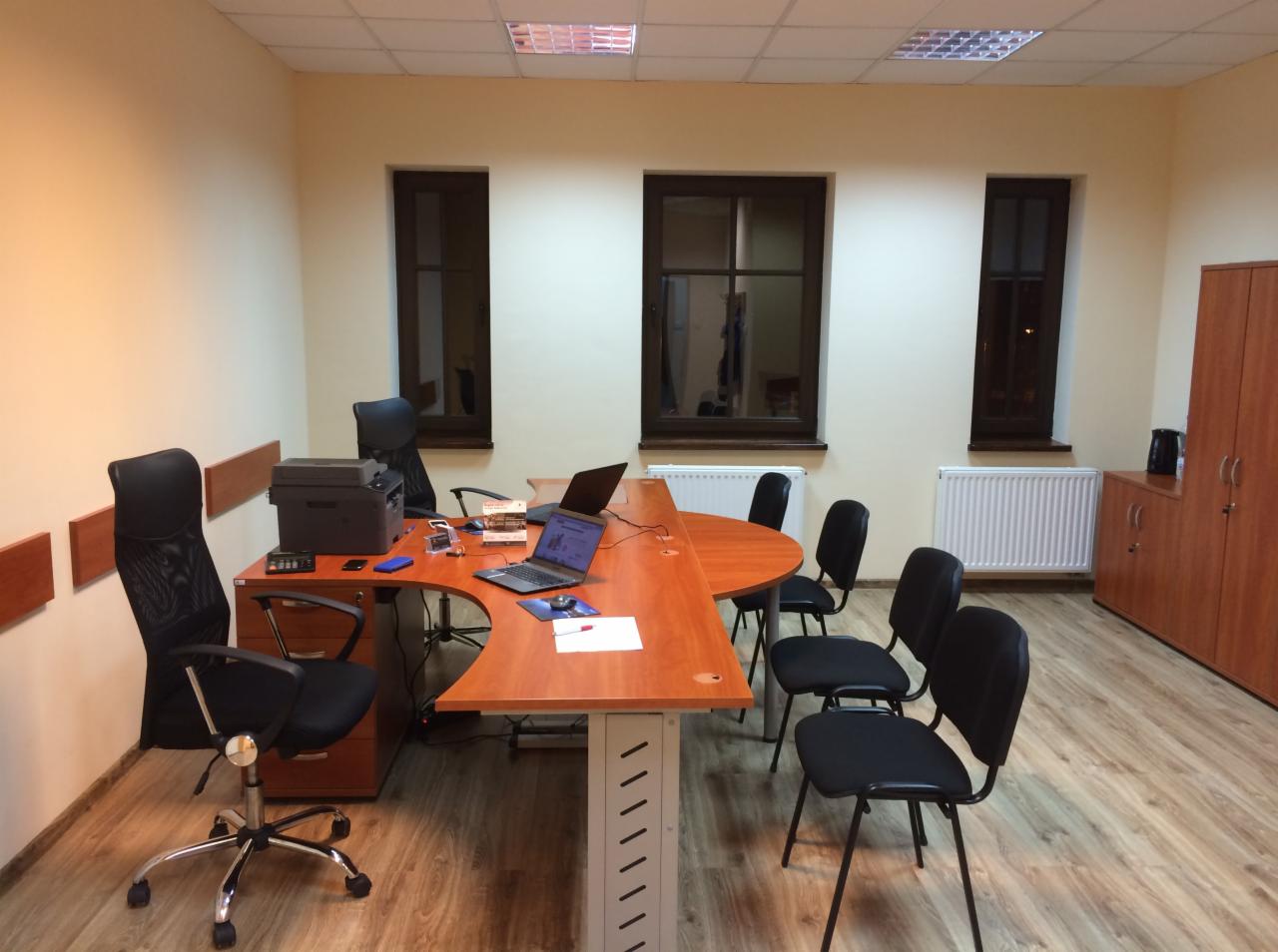 Biuro z dużym, zaokrąglonym stołem konferencyjnym, dwoma laptopami, drukarką i krzesłami biurowymi, oświetlone światłem dziennym wpadającym przez trzy okna.