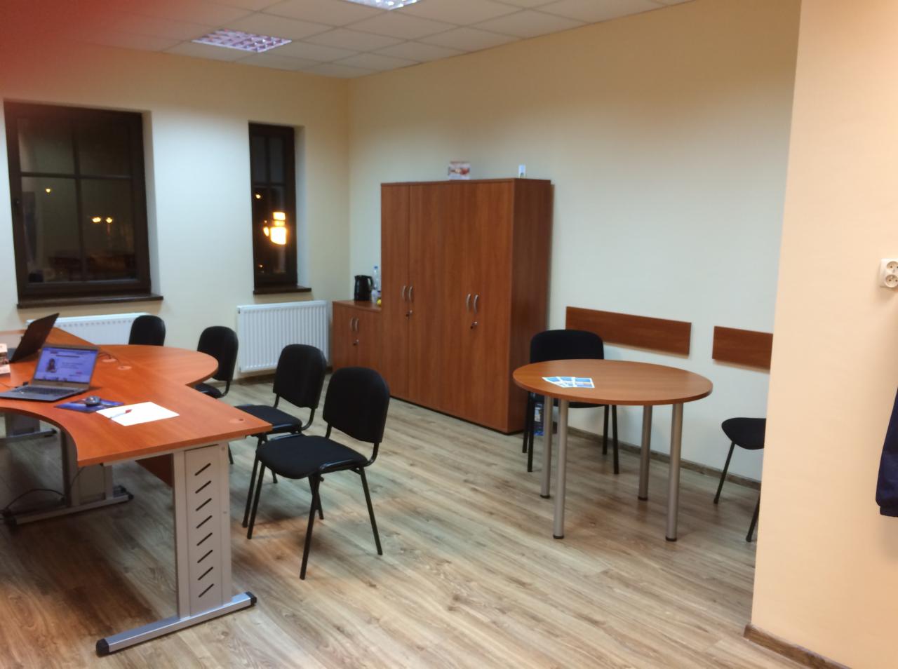 Jasne biuro z biurkami, krzesłami i szafą, widok na laptopa na biurku z boku okna.