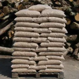 Spiętrzone worki z granulatem na palecie, w tle stos drewna opałowego.