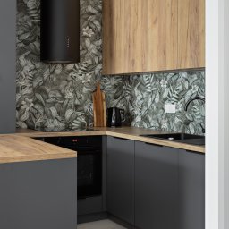 Anava Studio - Nowoczesna kuchnia w szarościach i drewnie: czarny okap cylindryczny, wzorzysta tapeta z motywem liści, drewniane blaty i górne szafki. Minimalistyczny design.