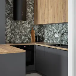 Nowoczesna kuchnia w szarościach i drewnie: czarny okap cylindryczny, wzorzysta tapeta z motywem liści, drewniane blaty i górne szafki. Minimalistyczny design.