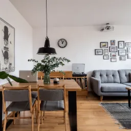 Jasne, przestronne wnętrze: drewniany stół z laptopem i wazonem, szara sofa z poduszkami, galeria zdjęć na ścianie, gramofon i dywan w stylu vintage.