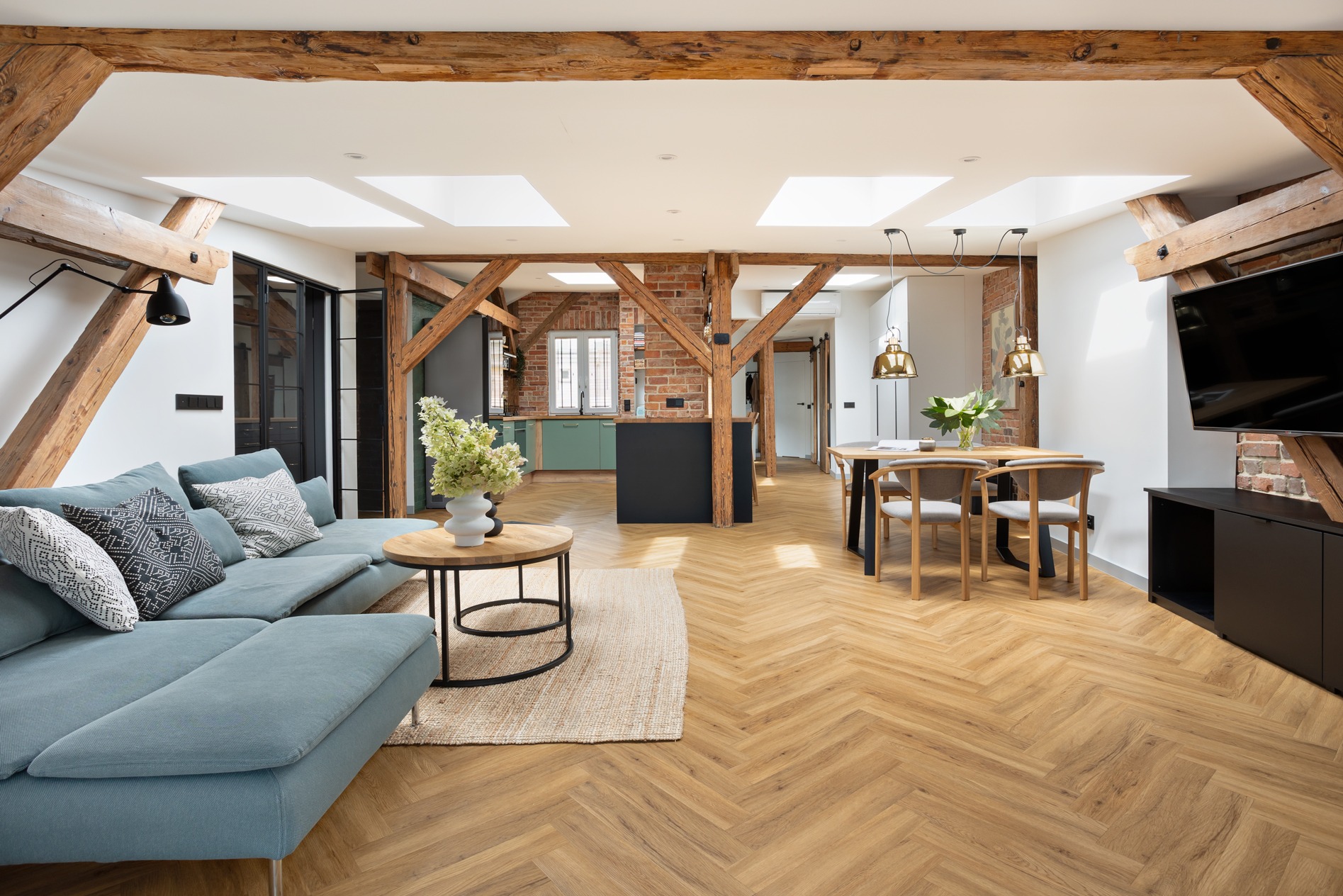 Jasne wnętrze z drewnianymi belkami stropowymi, błękitną sofą i parkietem w jodełkę. Widoczna kuchnia z zielonymi szafkami i ceglana ściana.