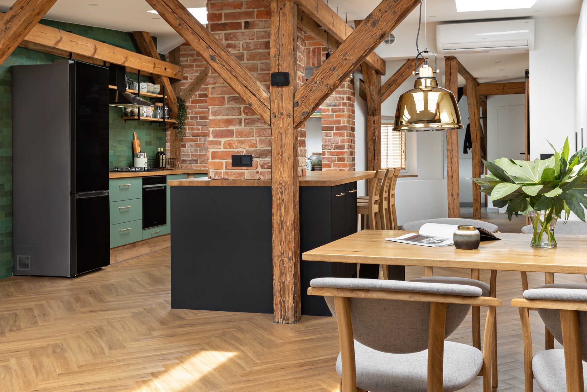 Jasna kuchnia z drewnianymi belkami stropowymi, zielonymi szafkami i czarną wyspą. Na pierwszym planie stół z szarymi krzesłami i wazonem z zielonymi liśćmi.