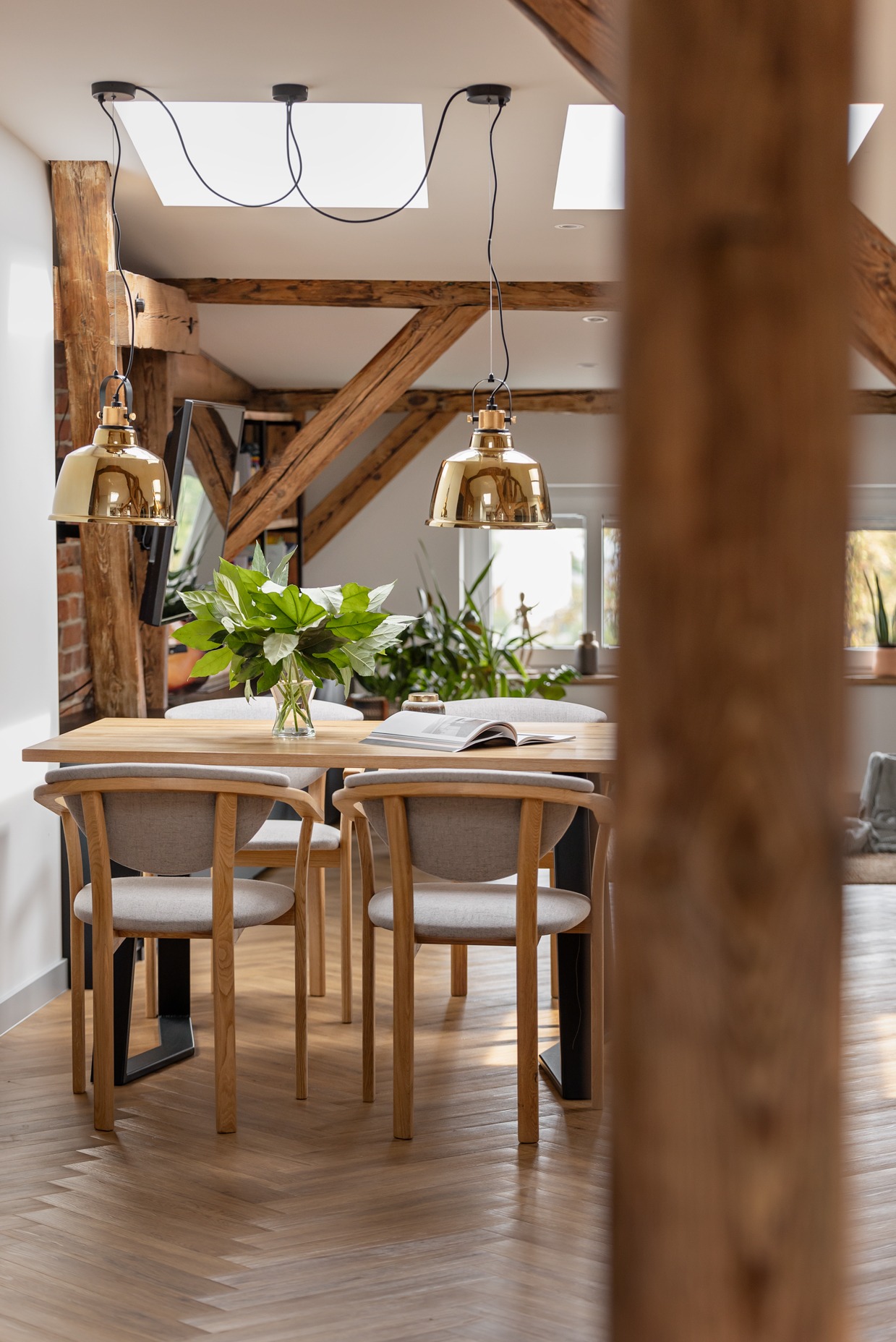 Jasna jadalnia w stylu skandynawskim: drewniany stół z bukietem, krzesła z szarą tapicerką, parkiet w jodełkę. Złote lampy wiszące pod skosami dachu z oknami.